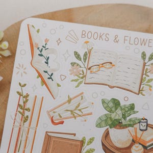 Peut inclure: Une planche d'autocollants avec le texte BOOKS & FLOWERS, illustr&eacute;e de livres ouverts, de piles de livres, de plantes en pot et de lunettes de lecture. Les motifs pr&eacute;sentent des tons doux marron, vert et orange avec des d&eacute;tails floraux.