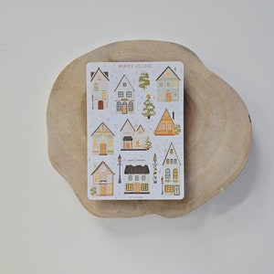 May include: A rectangular sticker sheet titled WINTER VILLAGE featuring various illustrated houses, trees, and street lamps in muted tones of brown, green, and cream. The designs include multiple architectural styles with windows, doors, and festive decorations.