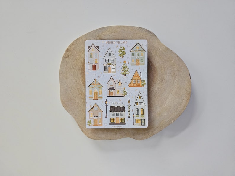 May include: A rectangular sticker sheet titled WINTER VILLAGE featuring various illustrated houses, trees, and street lamps in muted tones of brown, green, and cream. The designs include multiple architectural styles with windows, doors, and festive decorations.