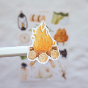 May include: A die-cut sticker featuring a campfire illustration with bright yellow and orange flames rising above a stack of brown logs. The sticker is held up against a blurred sticker sheet background that contains various other camping-themed icons.
