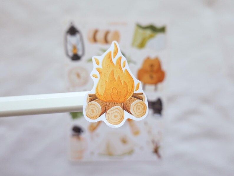 May include: A die-cut sticker featuring a campfire illustration with bright yellow and orange flames rising above a stack of brown logs. The sticker is held up against a blurred sticker sheet background that contains various other camping-themed icons.