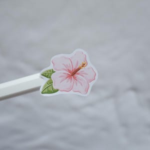 May include: A light pink hibiscus flower sticker with green leaves and a prominent stamen, attached to the end of a white pencil. The sticker features a white border around the floral illustration, showcasing soft pink petals and dark green foliage.