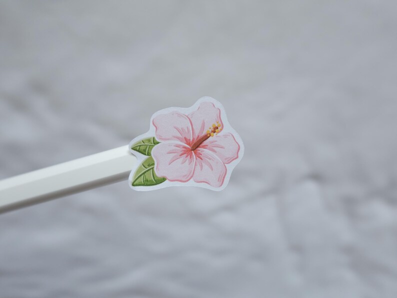May include: A light pink hibiscus flower sticker with green leaves and a prominent stamen, attached to the end of a white pencil. The sticker features a white border around the floral illustration, showcasing soft pink petals and dark green foliage.