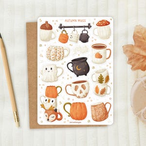 May include: A sticker sheet with illustrations of autumn mugs. Designs include a mushroom, pumpkin, ghost, and skull. The mugs are in shades of orange, brown, and white. The text "AUTUMN MUGS" is at the top. A pencil and a brown card are on the left.