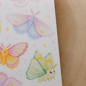 May include: A sticker sheet featuring multiple moth illustrations in pastel colors. The moths display pink, blue, yellow, and green wings with crescent moon patterns. Small decorative shapes like stars, circles, and triangles are scattered between the moths.