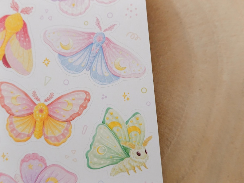 May include: A sticker sheet featuring multiple moth illustrations in pastel colors. The moths display pink, blue, yellow, and green wings with crescent moon patterns. Small decorative shapes like stars, circles, and triangles are scattered between the moths.