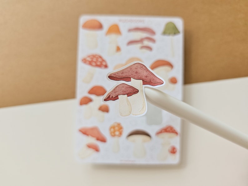 K&ouml;nnte beinhalten: Eine Bogen mit Aufklebern, die verschiedene Illustrationen von Pilzen in verschiedenen Farben und Stilen zeigen. Die Aufkleber sind auf wei&szlig;em Hintergrund und haben eine gl&auml;nzende Oberfl&auml;che.