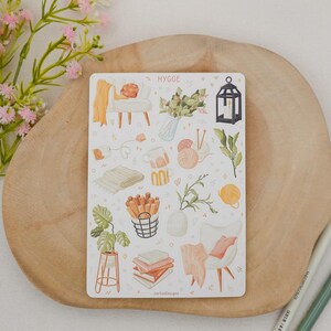 Op de afbeelding: Een stickervel met het woord "HYGGE" bevat illustraties van gezellige woondecoratie-items. De afbeeldingen omvatten een stoel, een deken, planten, boeken en een lantaarn. Het stickervel ligt op een houten oppervlak.