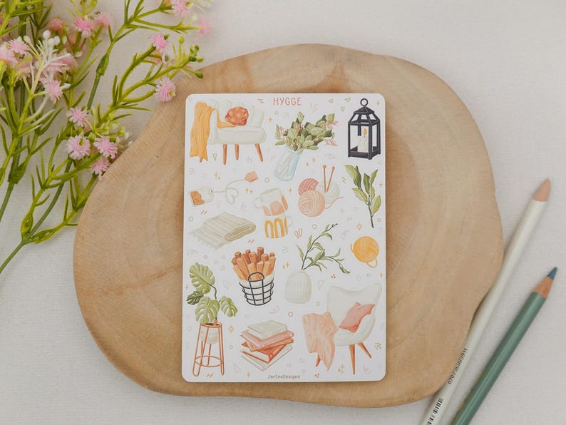 Op de afbeelding: Een stickervel met het woord "HYGGE" bevat illustraties van gezellige woondecoratie-items. De afbeeldingen omvatten een stoel, een deken, planten, boeken en een lantaarn. Het stickervel ligt op een houten oppervlak.