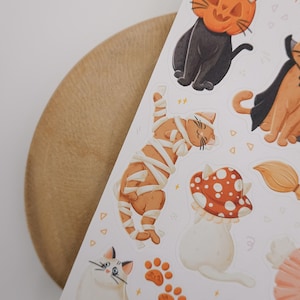 Pu&ograve; includere: Un foglio di adesivi con varie illustrazioni di gatti a tema Halloween, tra cui un gatto nero con testa di zucca, un gatto arancione con mantello da vampiro, un gatto arancione avvolto in bende da mummia e un gatto bianco sotto un fungo.