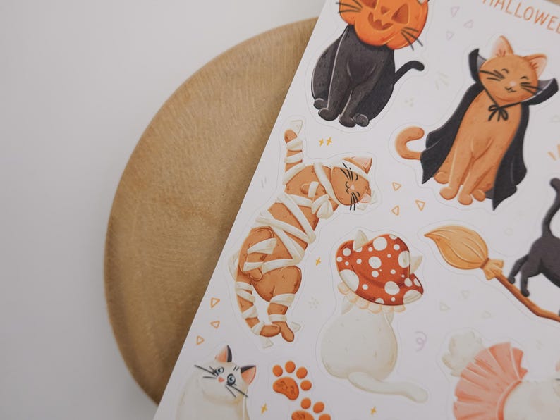 Pu&ograve; includere: Un foglio di adesivi con varie illustrazioni di gatti a tema Halloween, tra cui un gatto nero con testa di zucca, un gatto arancione con mantello da vampiro, un gatto arancione avvolto in bende da mummia e un gatto bianco sotto un fungo.