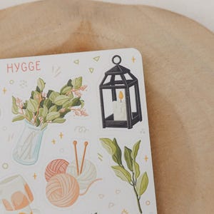 Op de afbeelding: Een stickervel met een gezellig thema, met illustraties van een zwarte lantaarn met een brandende kaars, een vaas met groen, bollen wol en een mok. Het woord "HYGGE" is in oranje geschreven.