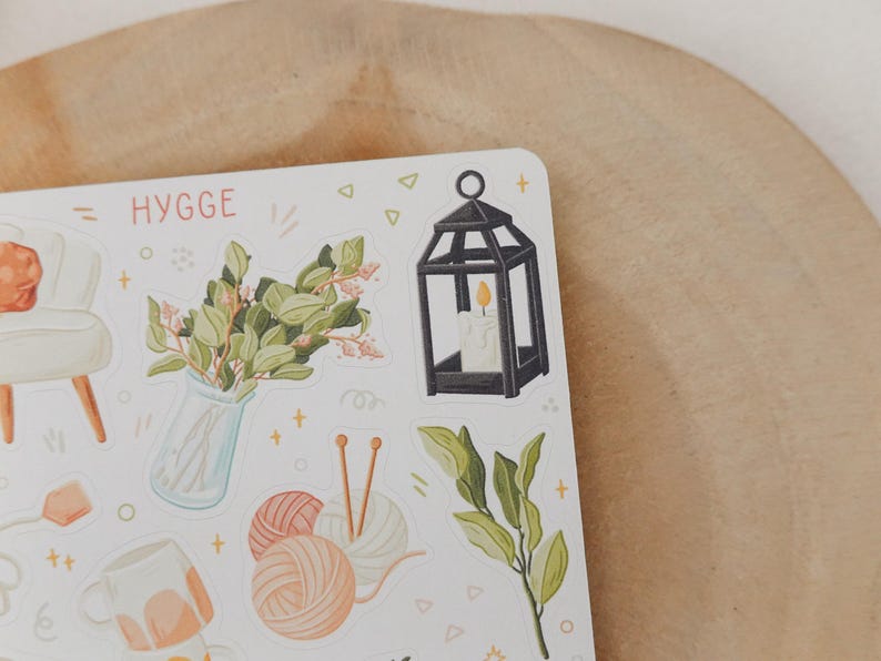 Op de afbeelding: Een stickervel met een gezellig thema, met illustraties van een zwarte lantaarn met een brandende kaars, een vaas met groen, bollen wol en een mok. Het woord "HYGGE" is in oranje geschreven.