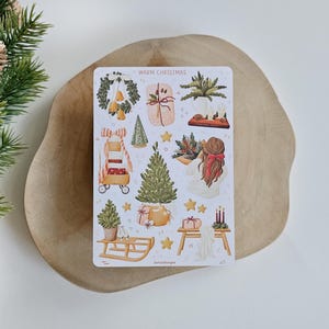 May include: A rectangular sticker sheet titled WARM CHRISTMAS featuring various holiday illustrations including a wreath, wrapped gifts, a Christmas tree, a wooden sled, a person with a red hair bow, and candles, all rendered in a soft, watercolor style.