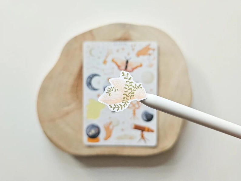Puede incluir: Una pegatina troquelada que muestra un cristal rosa p&aacute;lido envuelto en enredaderas verdes, sostenida por un l&aacute;piz &oacute;ptico blanco. Al fondo, una hoja de pegatinas rectangular muestra varias ilustraciones, incluyendo una luna creciente, una polilla y un telescopio.