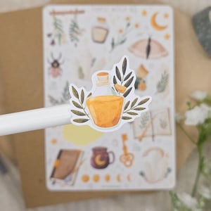 Puede incluir: Una hoja de pegatinas con varias ilustraciones que incluyen una botella de poci&oacute;n, un escarabajo, una luna creciente, una llave y libros. Una pegatina de botella con l&iacute;quido amarillo y hojas verdes se sostiene en primer plano sobre un bol&iacute;grafo.
