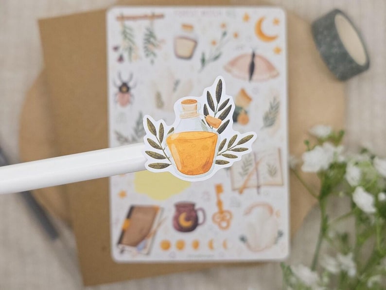 Puede incluir: Una hoja de pegatinas con varias ilustraciones que incluyen una botella de poci&oacute;n, un escarabajo, una luna creciente, una llave y libros. Una pegatina de botella con l&iacute;quido amarillo y hojas verdes se sostiene en primer plano sobre un bol&iacute;grafo.