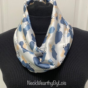 May include: A white satin infinity scarf with a blue and beige abstract print. The scarf is on a black mock neck shirt. The text "NeckWearthyByLois" is visible at the bottom of the image.