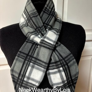 May include: A gray, white, and black plaid fleece scarf. The scarf is draped over a black form, showcasing the pattern and stitching details. The text "NeckWearthyByLois" is visible at the bottom of the scarf.