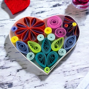 May include: A white heart-shaped frame filled with colorful paper rolls in a variety of shapes and sizes, creating a floral design.