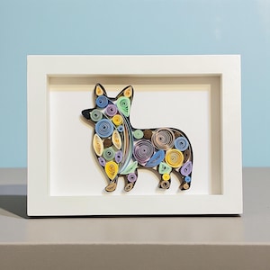 May include: A framed paper quilling art piece featuring a colorful corgi dog. The corgi is made of tightly rolled paper strips in shades of yellow, blue, purple, and brown. The artwork is set against a white background within a white frame.