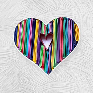 May include: A colorful heart-shaped paper craft made of layered strips of paper in various shades of red, orange, yellow, green, blue, purple, and pink. The heart is outlined in white and has a smaller pink heart in the center.