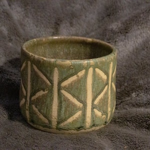 Może przedstawiać: Zielona doniczka ceramiczna z białym geometrycznym wzorem. Doniczka jest wykonana z ceramiki i ma rustykalny, ręcznie robiony wygląd.