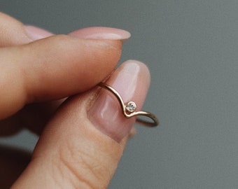 May include: A delicate gold ring with a small diamond set in the centre. The ring is shaped like a V and is worn on a finger.