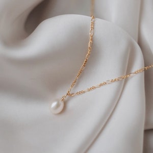 May include: A delicate gold chain necklace with a single white pearl pendant. The necklace is displayed on a white fabric background.