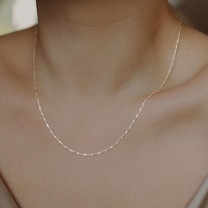 Op de afbeelding: Fijne gouden ketting met een subtiele, getextureerde ketting. De ketting wordt gedragen door iemand in een satijnen top. De sieraden zijn een eenvoudig, elegant accessoire.