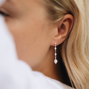 Könnte beinhalten: Ein Paar silberne Ohrringe mit kleinen weißen Perlen. Die Ohrringe hängen am Ohrläppchen einer Person mit blonden Haaren.