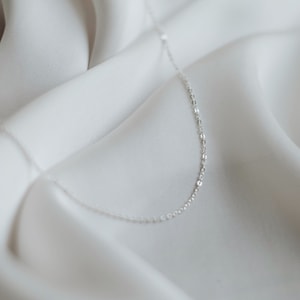 May include: A delicate silver chain necklace on a white fabric background.