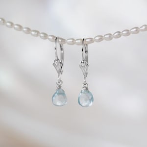 Op de afbeelding: Een paar zilveren oorhangers met lichtblauwe, druppelvormige edelstenen. De oorbellen hangen aan een delicate parelketting tegen een zachte, witte achtergrond. De oorbellen hebben een decoratief ontwerp boven de edelsteen.