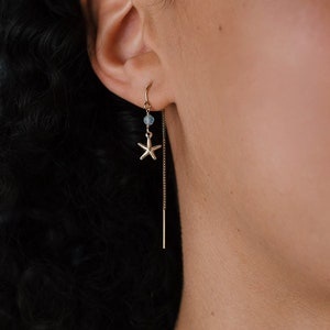 Op de afbeelding: Een gouden kettingoorring met een kleine blauwe kraal en een zeester-bedel. De oorring wordt gedragen door een persoon met donker krullend haar.