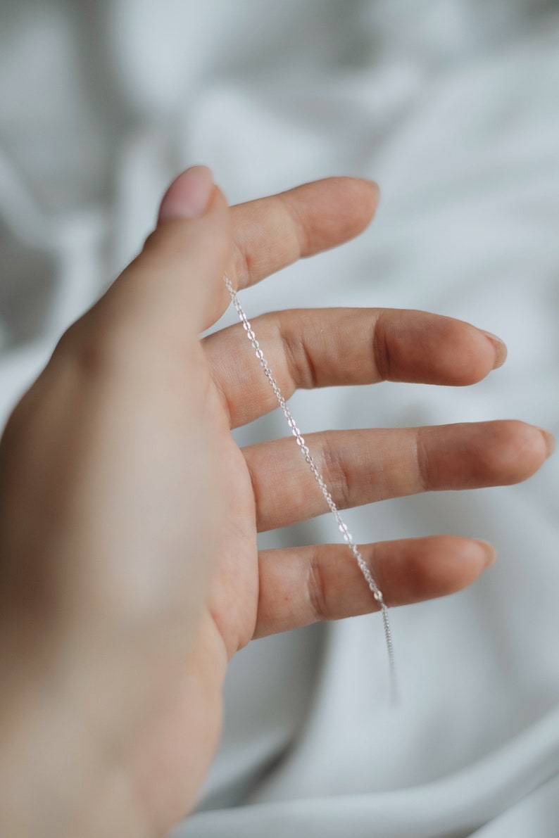 May include: A silver chain necklace, held in a hand, against a white background.