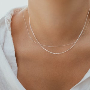 May include: A close-up of a person wearing two delicate silver necklaces. The necklaces are layered, with one being a thin chain and the other a slightly thicker chain with small, round links. The person is wearing a white shirt.
