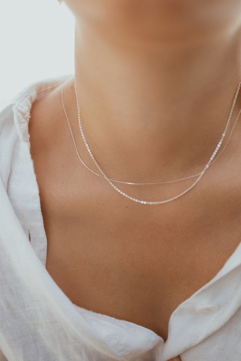 May include: A close-up of a person wearing two delicate silver necklaces. The necklaces are layered, with one being a thin chain and the other a slightly thicker chain with small, round links. The person is wearing a white shirt.