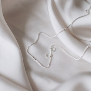 May include: A silver chain necklace on a white fabric background.