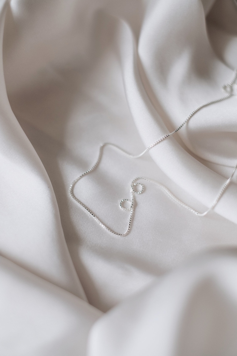 May include: A silver chain necklace on a white fabric background.