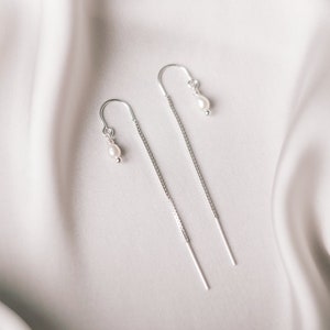 Op de afbeelding: Een paar zilveren kettingoorbellen met kleine parel accenten. De oorbellen hangen aan een witte stoffen achtergrond.