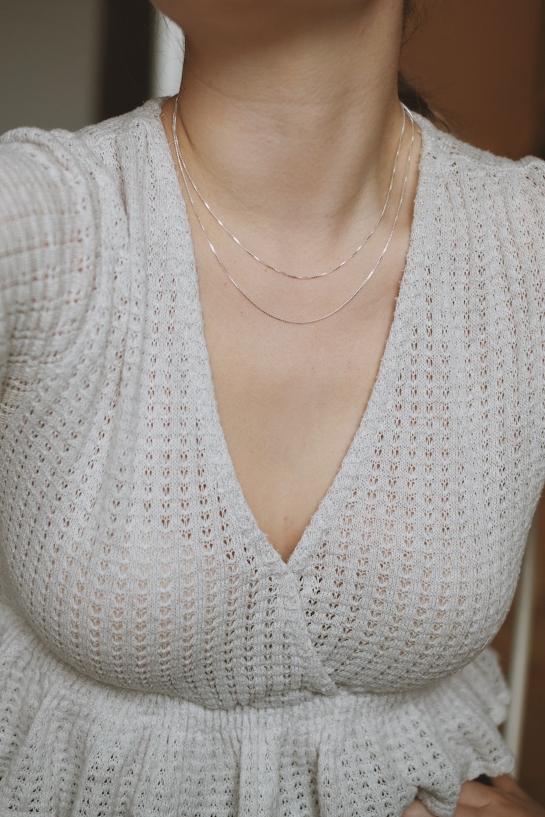 May include: A close-up of a person wearing a white, crocheted, v-neck top with a silver chain necklace.