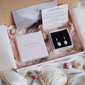 May include: A pink gift box with the words "Sophistiqu&eacute; Jewellery" printed on the lid. The box is open and contains a smaller white box with a pair of pearl drop earrings. The earrings are gold and have a delicate design. The box is surrounded by white tissue paper and pink flowers.
