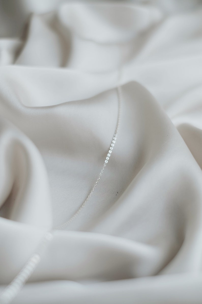 May include: A delicate silver chain necklace on a white fabric background.