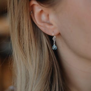 Op de afbeelding: Een zilveren oorbel met een beugel sluiting en een klein, sierlijk ontwerp. De oorbel heeft een druppelvormige, lichtblauwe edelsteen. De afbeelding toont de oorbel gedragen aan een oor, met blond haar op de achtergrond.