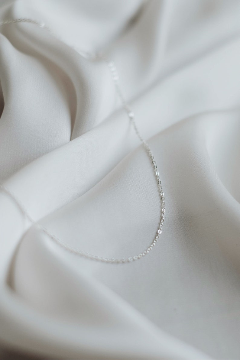 May include: A delicate silver chain necklace on a white fabric background.
