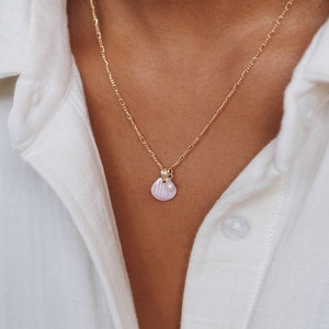 Puede incluir: Un collar de cadena dorada con un delicado colgante de concha de mar y un pequeño acento de perla. El collar lo lleva una persona que lleva una camisa blanca.