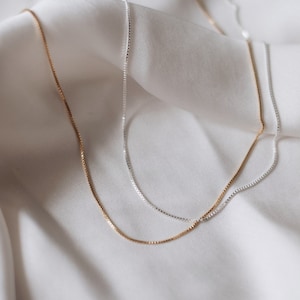 May include: Two delicate chain necklaces, one silver and one gold, lay on a white fabric background. The chains are both box chains, with a simple and elegant design.