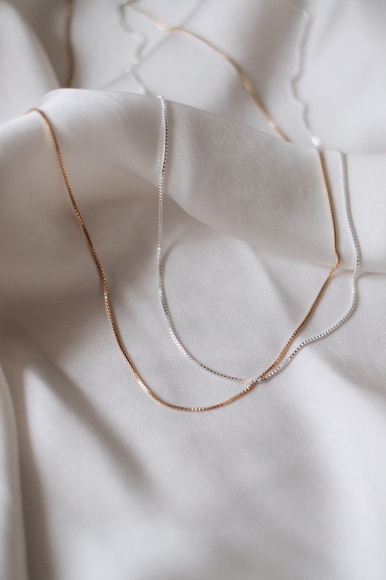 May include: Two delicate chain necklaces, one silver and one gold, lay on a white fabric background. The chains are both box chains, with a simple and elegant design.