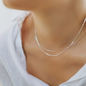 May include: A close-up of a person's neck wearing two delicate silver necklaces. The necklaces are layered, with one being a thin chain and the other a slightly thicker chain. The person is wearing a white shirt.