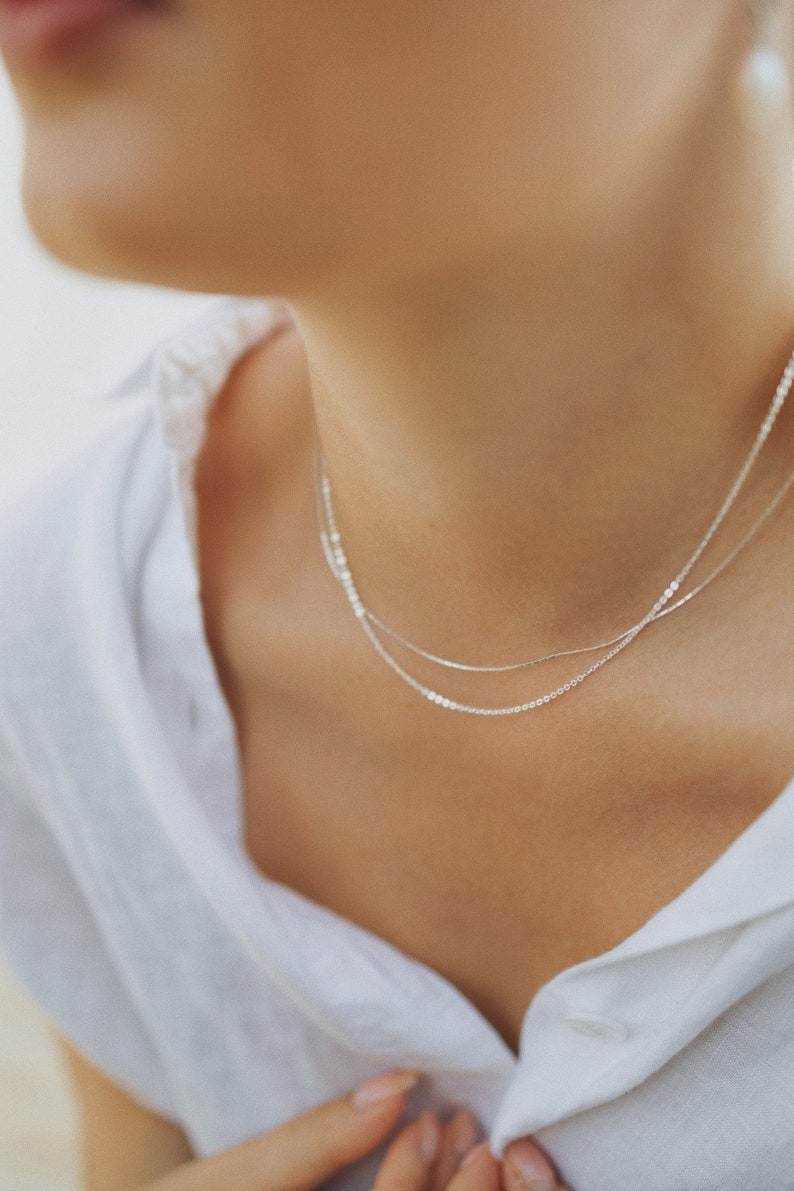 May include: A close-up of a person's neck wearing two delicate silver necklaces. The necklaces are layered, with one being a thin chain and the other a slightly thicker chain. The person is wearing a white shirt.